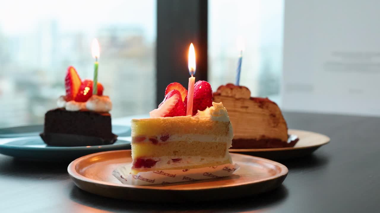 Three assorted cakes with lit candles on plates, natural daylight, shallow depth of field