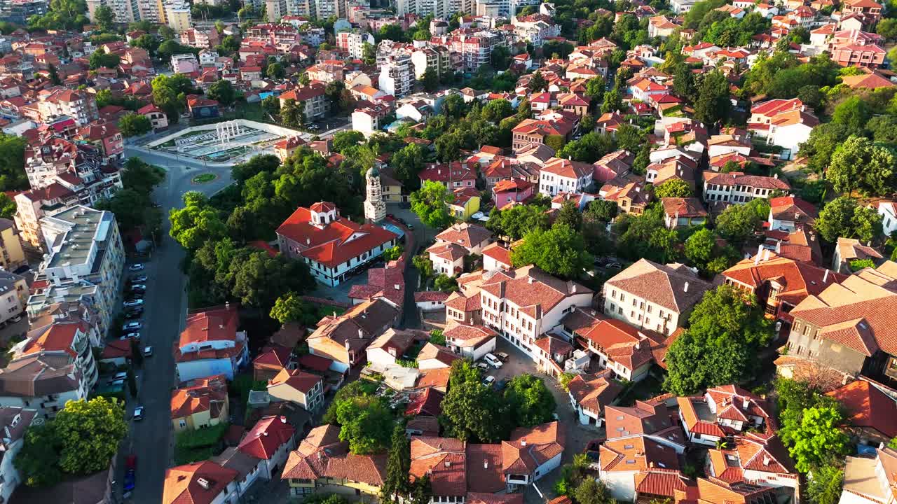 O drone percorre o terreno variado de Plovdiv, destacando telhados, vegetação e blocos de apartamentos em uma luz suave e quente.