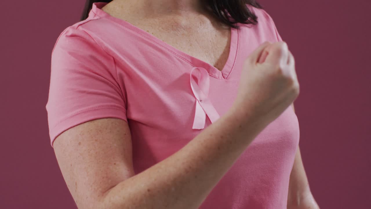 ピンクの背景を背に、胸にピンクリボンをつけ拳を握りしめている女性の胴体部分
