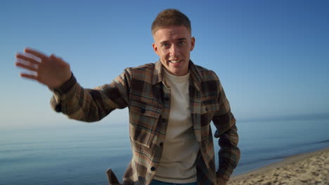 Attractive-man-waving-hand-on-camera-standing-beach.-Stylish-handsome-posing.