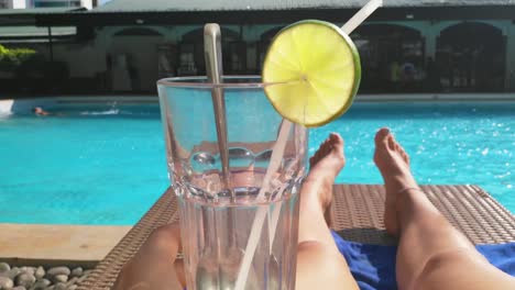 first person view of hand holding drink with slice of lime and straw at poolside in beautiful sunny day.