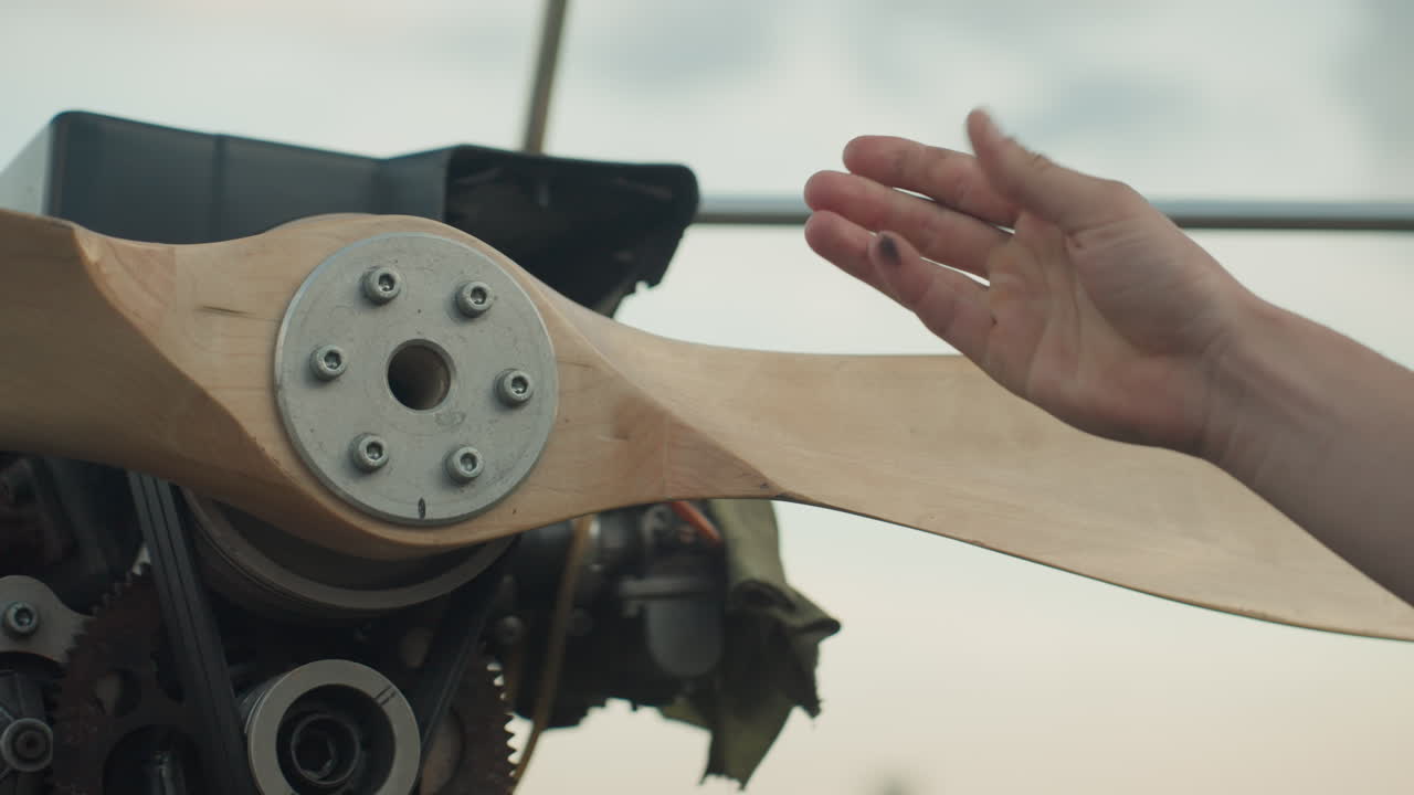 close up of hand wiping wooden fan blade near metal frame in airy outdoor setting with soft sky backdrop, focus on gentle cleaning motion and textured grain under natural light