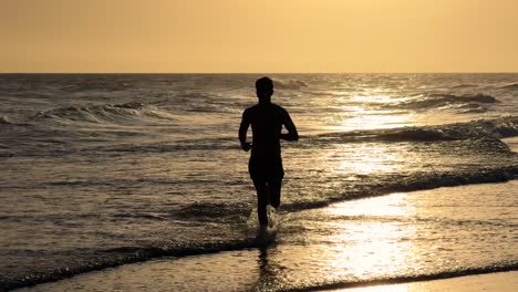 Junger-Mann-Läuft-Bei-Sonnenuntergang-Am-Strand-Entlang-In-Richtung-Kamera