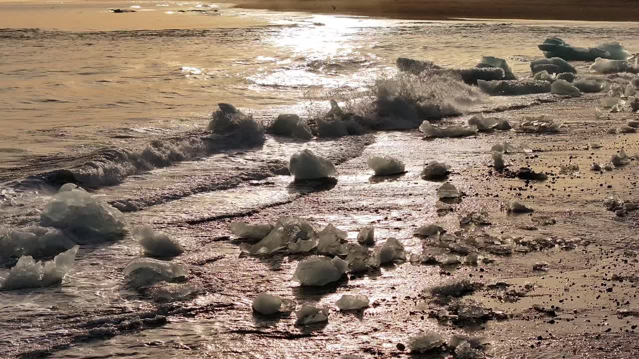 Waves crash on Diamond Beach in Iceland, scattering glistening ice chunks across the black sand as the golden light of sunset reflects on the water.
