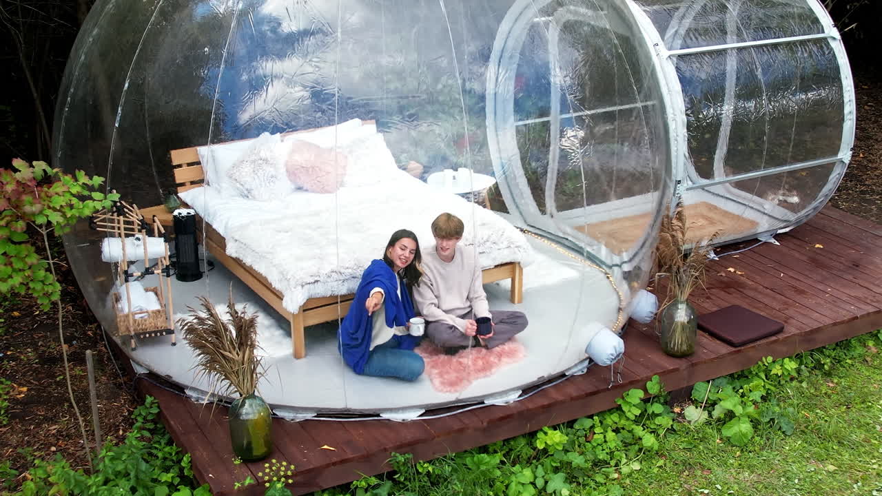 Gennemsigtigt bobletelt ved glamping, frodig skov rundt om og indvendigt. Et par sidder under et tæppe, holder kopper og snakker