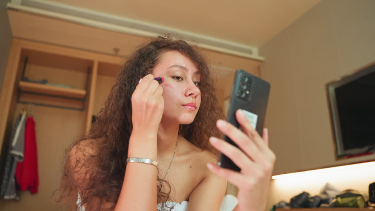 Hotel room guest applies makeup using phone. Warm bedside lighting, wooden wardrobe, wallmounted TV, towelwrapped hair, mirror selfie angle, blending concealer and powder, relaxed evening preparation