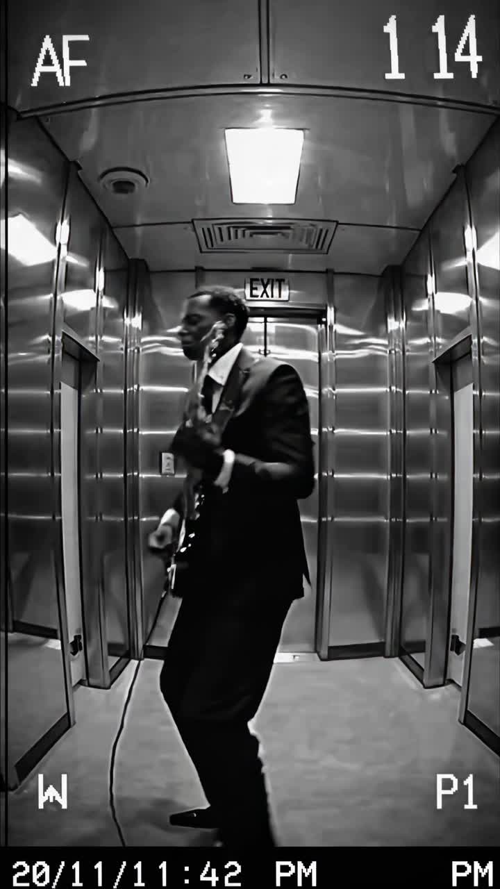 A black-and-white video still of a man playing guitar in an elevator, captured from a low-angle