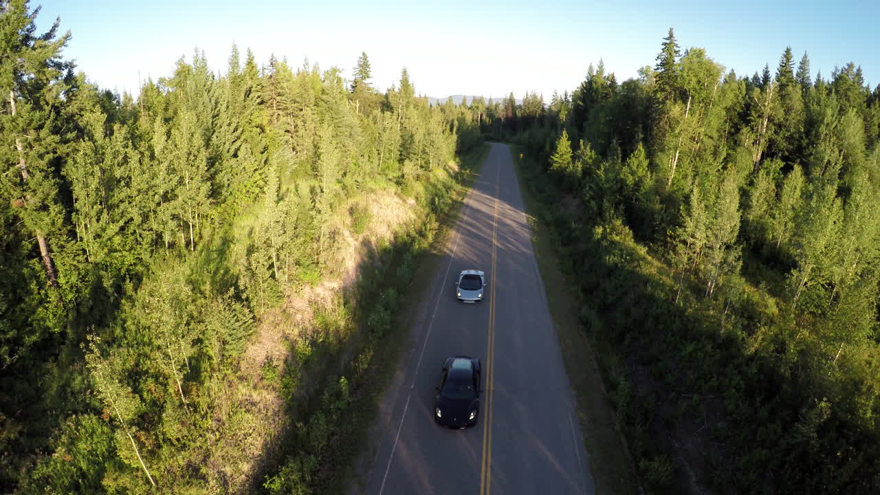 Sports cars cruise down a country road in the remote wilderness, as viewed from a drone overhead nearing sunset.