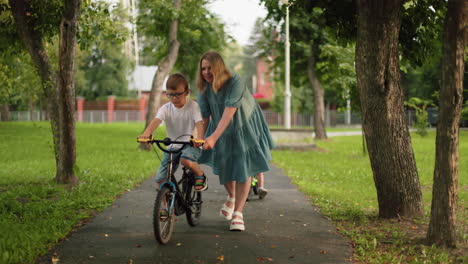 una madre apoya a su hijo mientras anda en bicicleta por un camino del parque, guiándolo mientras el niño más pequeño lo sigue