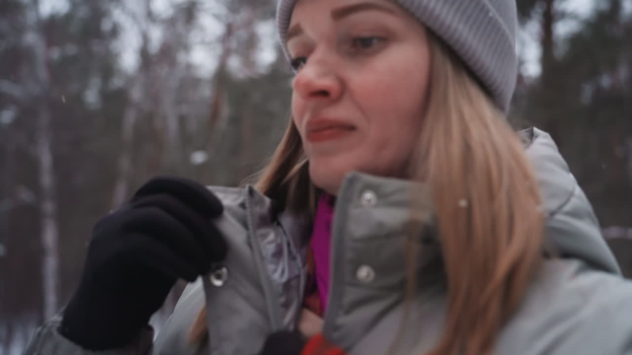 Nainen harmaassa talvitakissa ja mustissa hanskoissa seisoo lumisessa metsässä säätäen vetoketjua pitäen samalla punaista hihnaa, osittain epätarkkana, kuvaten kerroksellista ulkoiluvaatetusta ja liikettä kylmällä säällä.
