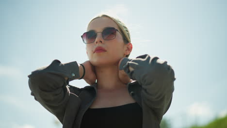 woman in black top under grey jacket wearing sunglasses, adjusting her hair and touching her collar with a focused expression, she stands outdoors under a bright sunny sky