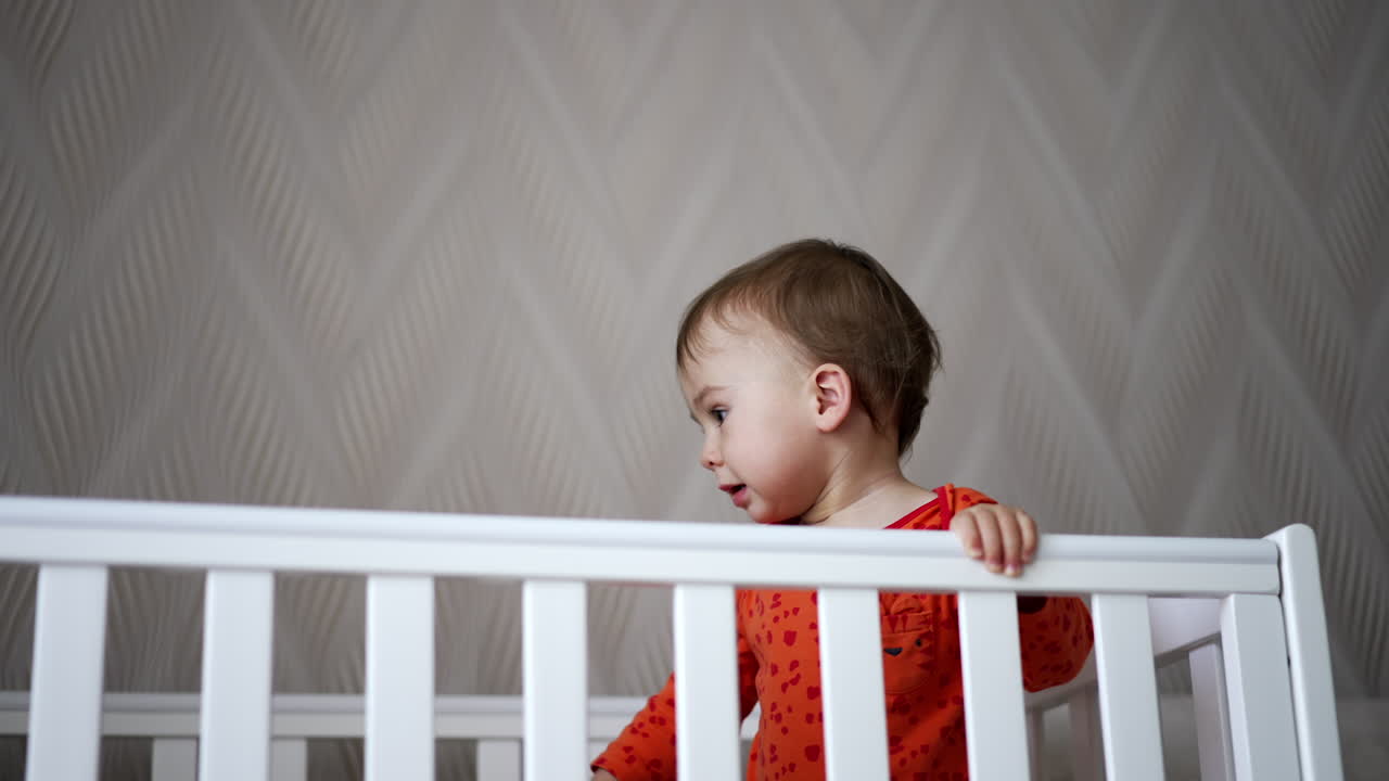 Calm cute baby in orange shirt standing in the cot. Beautiful child making sounds, holding a pacifier and looking at camera.