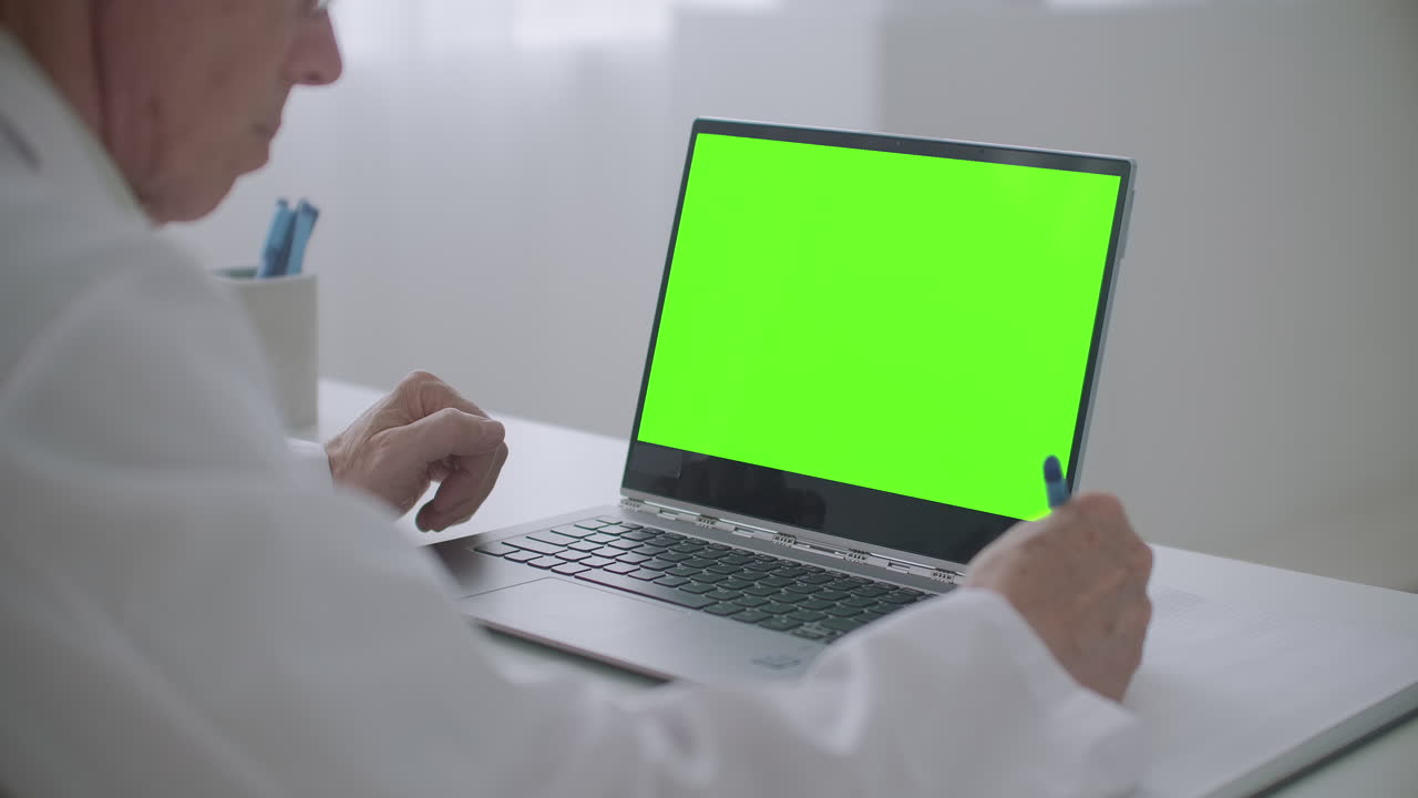 el médico está mirando la computadora portátil con pantalla de teclado cromático en la mesa en la oficina de la clínica escribiendo notas