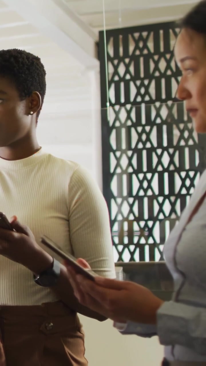 Vertical video: Two office women scrolling tablet, left tapping, HUD hexagons showing business data