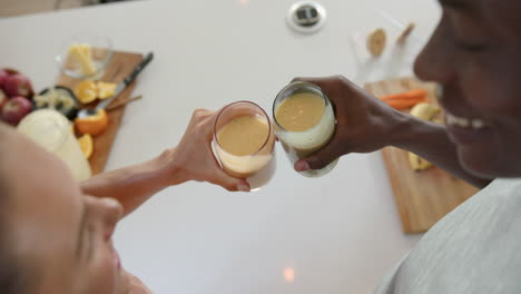 holding glasses of smoothies, diverse couple toasting in kitchen with fresh ingredients