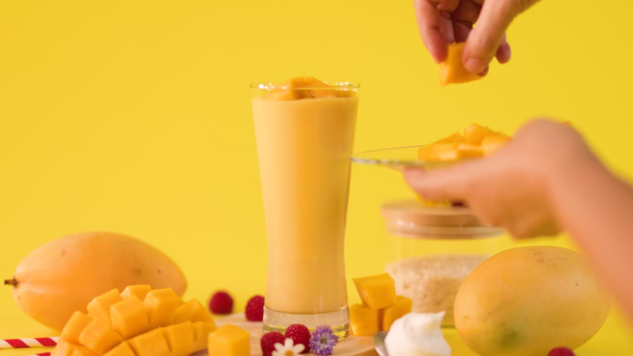 Human hand adds mango cubes to tall smoothie glass on bright yellow studio background