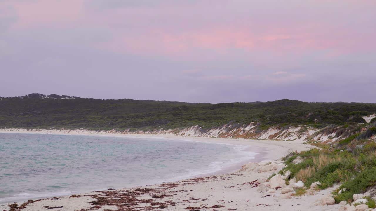 A serene white sandy beach with a soft purple glow in the clouds from the setting sun, creating a peaceful, magical atmosphere as the day transitions into evening.