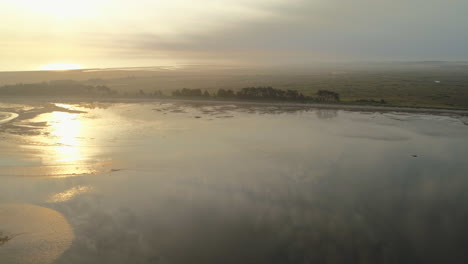 High-Establishing-Drone-Shot-Over-Still-Water-Towards-Sandy-Beach-with-Trees-with-Salt-Marsh-Fields-Behind-at-Stunning-Sunrise-in-Wells-Next-The-Sea-North-Norfolk-UK-East-Coast