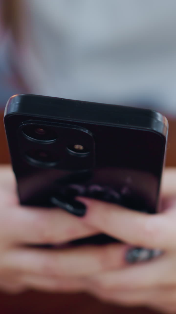 vista borrosa de la mano de la dama con uñas pulidas negras que se involucran con el teléfono inteligente colocado en una mesa de madera, rodeando el fondo borroso con objetos oscuros verdes y rosados