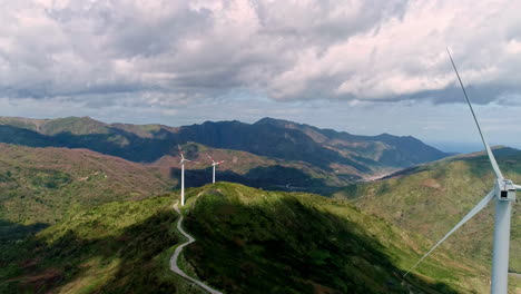 aerial view of wind turbines on path between green mountains during cloudy day - production of renewable energy in nature