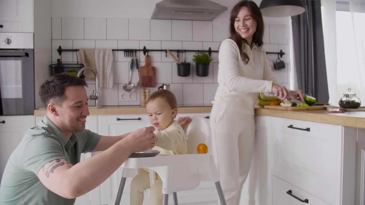 Happy Father Feeding His Cute Baby Girl Sitting In Her High Chair In The Kitchen While Smiling Mother Cutting Fruit And Looking At Them