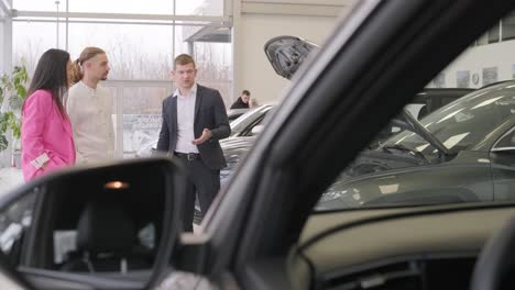 young car salesman showing to young couple new automobile at dealership salon.