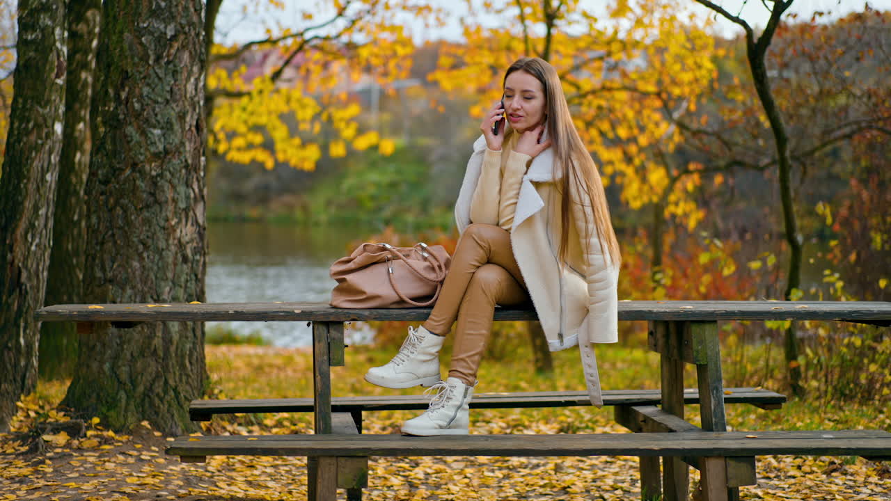 Attractive long-haired brunette having phone conversation outdoors. Lady in warm outfit sitting on the table in the beautiful autumn nature.