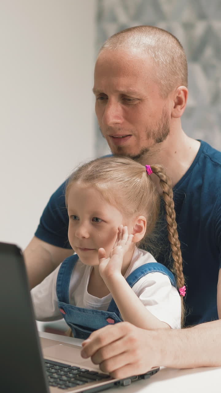 bearded dad and young daughter with long hair and pigtails sits on his lap, communicate with friends via laptop and video conference, wave his hands hello, smile, point his finger at monitor