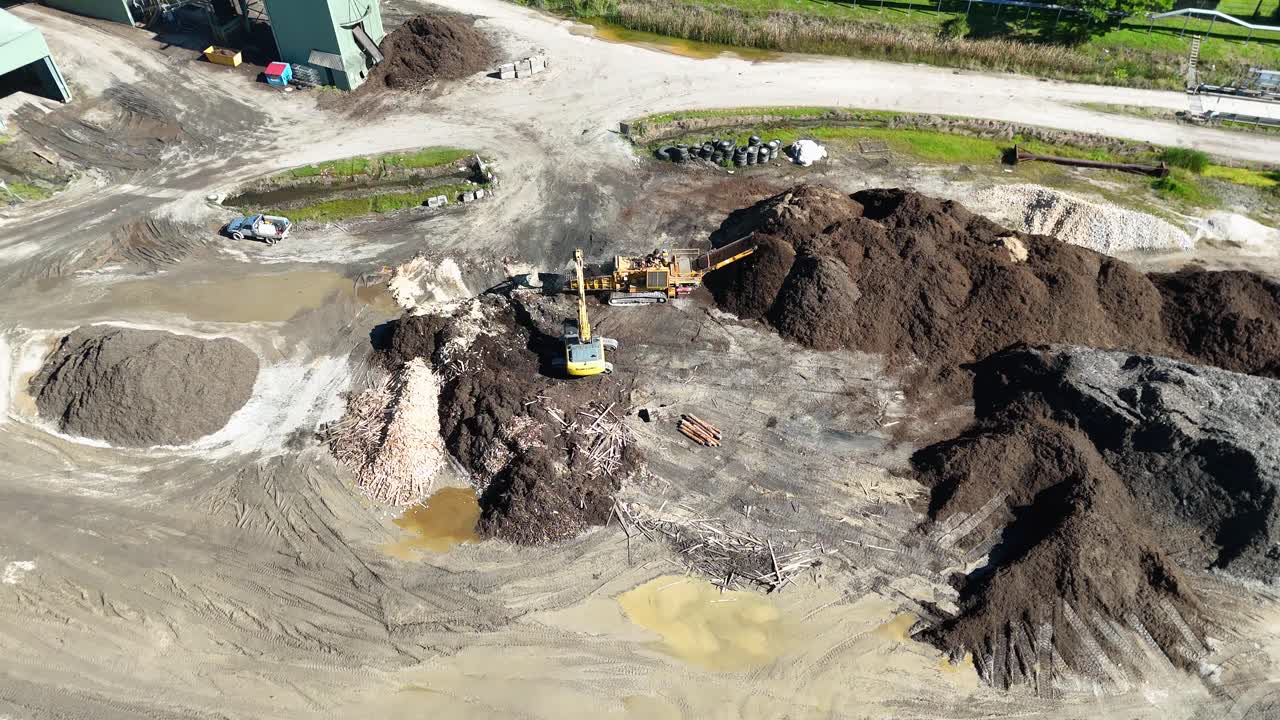 Drone footage captures machinery processing soil and mulch at an industrial site under bright daylight
