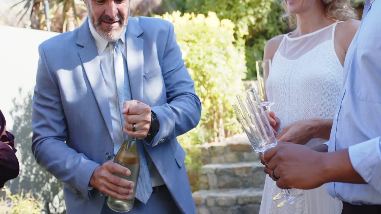 Gruppo eterogeneo che guarda l'ospite in abito blu stappare lo spumante sul patio, mentre tiene in mano dei flute e brinda.