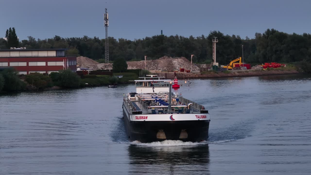 A cargo ship sails through a calm industrial canal at dusk with a backdrop of construction sites