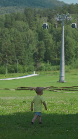 adorable little boy walks along green meadow of forest clearing under contemporary ropeway spending time in mountain park on summer day slow motion