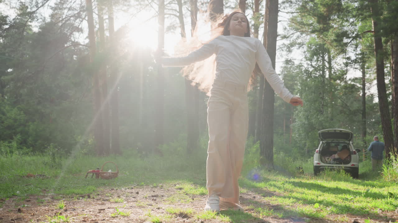 Teenage girl performing cartwheel on picnic mat in sunny forest clearing near parked family car, surrounded by green trees and soft sunlight, expressing joy, and freedom in peaceful summer nature