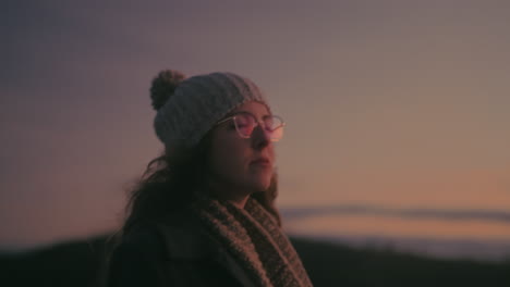 young woman in glasses, beanie and gray coat, walks on a portuguese mountain road, admiring picturesque sunset view. cinematic shot promotes revitalizing power of nature and escape from daily routines