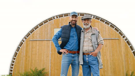 caucasian young man and his senior father smiling and looking at camera outside the stable in the field