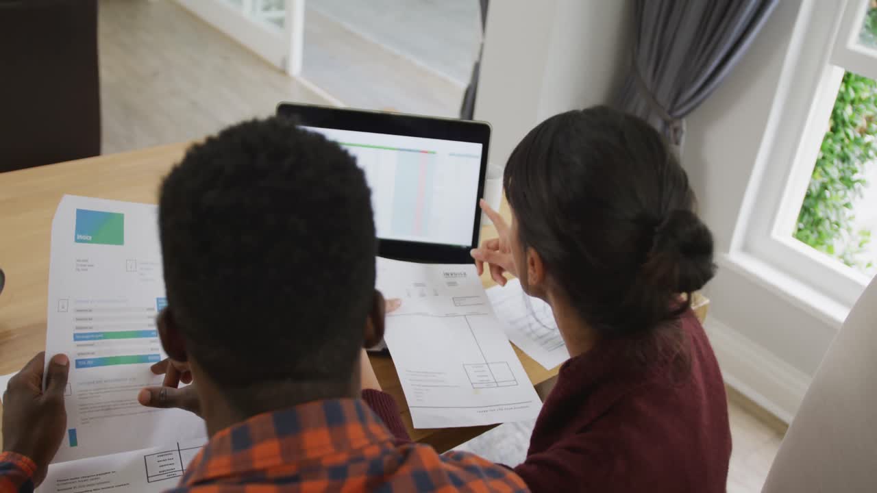 Diverse couple sitting at table talking and working with laptop