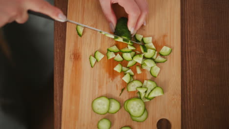 top down view of person slicing cucumber into small pieces on wooden cutting board, with scattered cucumber slices spread across surface during meal preparation in kitchen