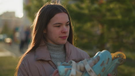 una mujer joven con una chaqueta de melocotón y un suéter de cuello gordo gris está de pie en un parque iluminado por el sol, sosteniendo un par de patines azules con una leve sonrisa en su cara