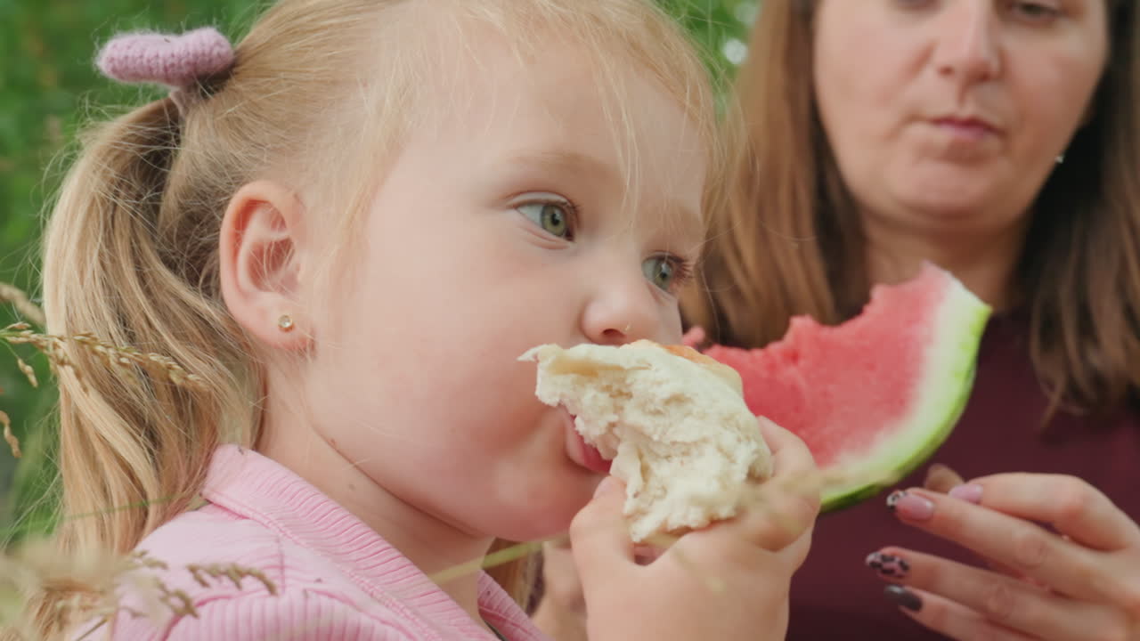 white child eating watermelon and bread, sticky fingers, crumbs on chin, mother serving slice, woodland picnic backdrop, closeup facial expression showing contentment and curiosity, summer warmth