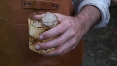 man in leather apron holds a whiskey glass with ice