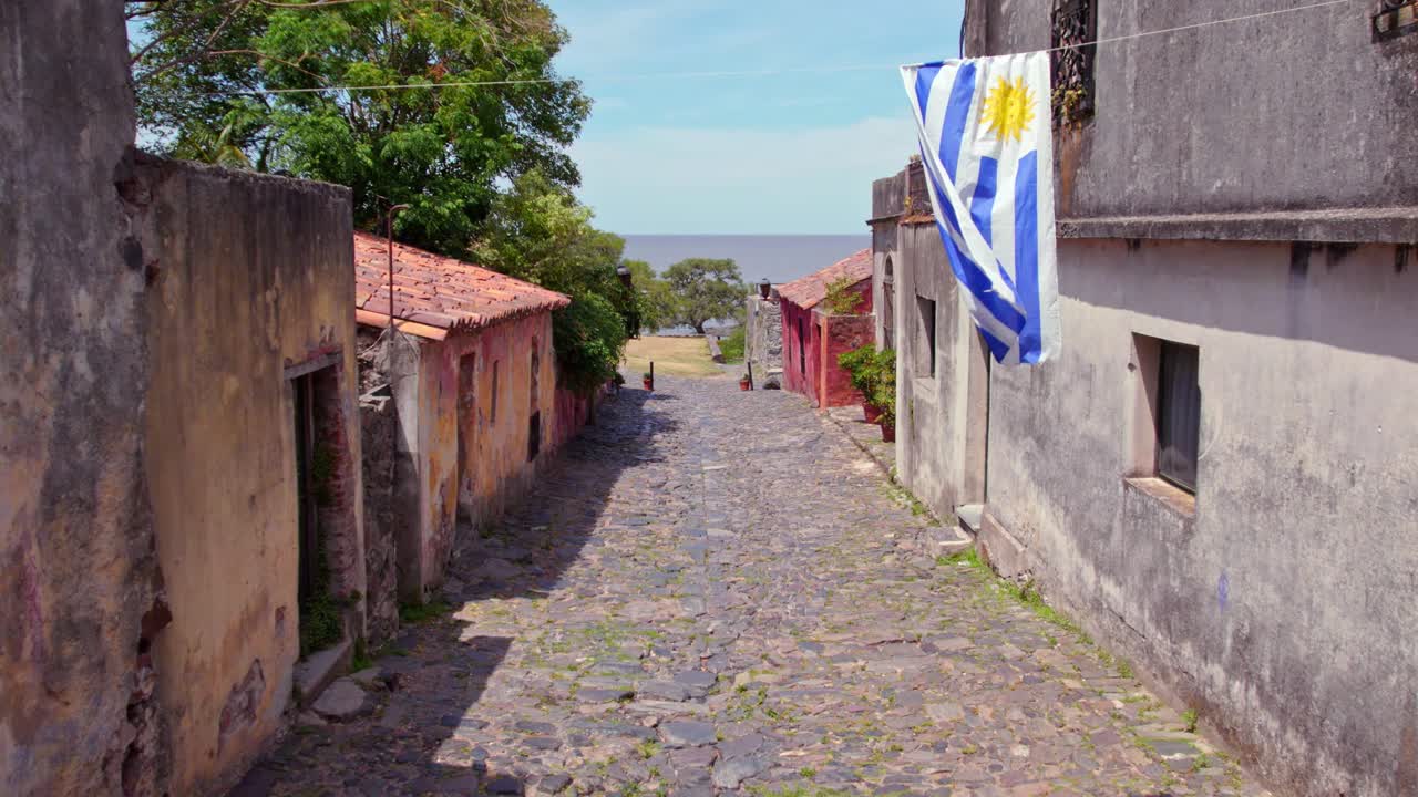 vista panorámica del carro desde la calle de los suspiros en el casco antiguo de colonia del sacramento, bandera de uruguay ondeando en cámara lenta