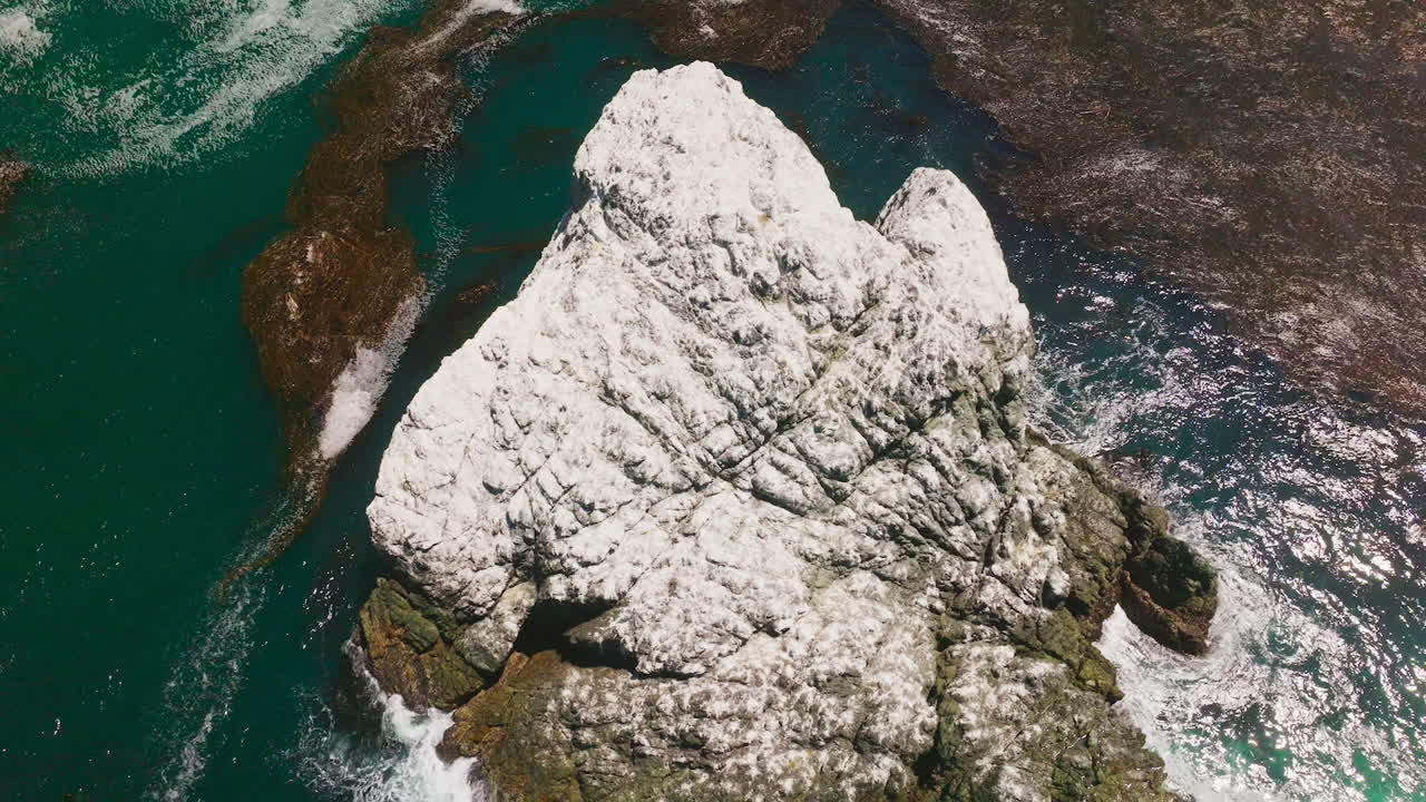 White rock peeping out from blue water of ocean on sunny day. Waves hitting the mount. Seaweeds floating on the surface of water. Top view.