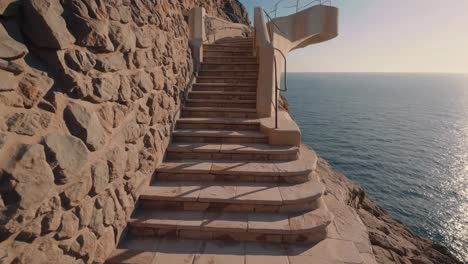 sunlit cliffside stairs descending towards the calm mediterranean sea at sunset, creating a scenic view with a stone wall on one side and a metal railing on the other