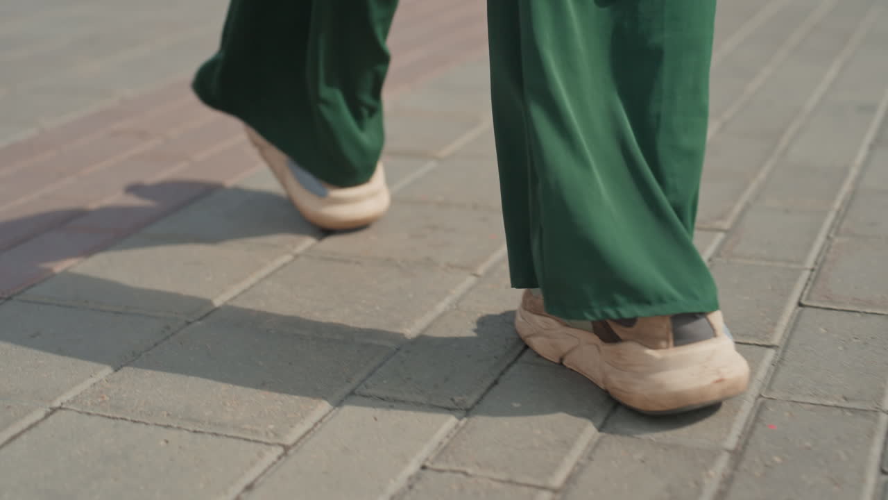 sunlit closeup footsteps on city pavement showing beige sneakers and green pants, rhythmic stride across textured tiles, long shadows and steady pace suggesting commuter routine through downtown