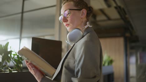 mujer de negocios rubia de mediana edad con gafas en un uniforme de negocios gris se encuentra en la oficina y mira un libro mientras trabaja