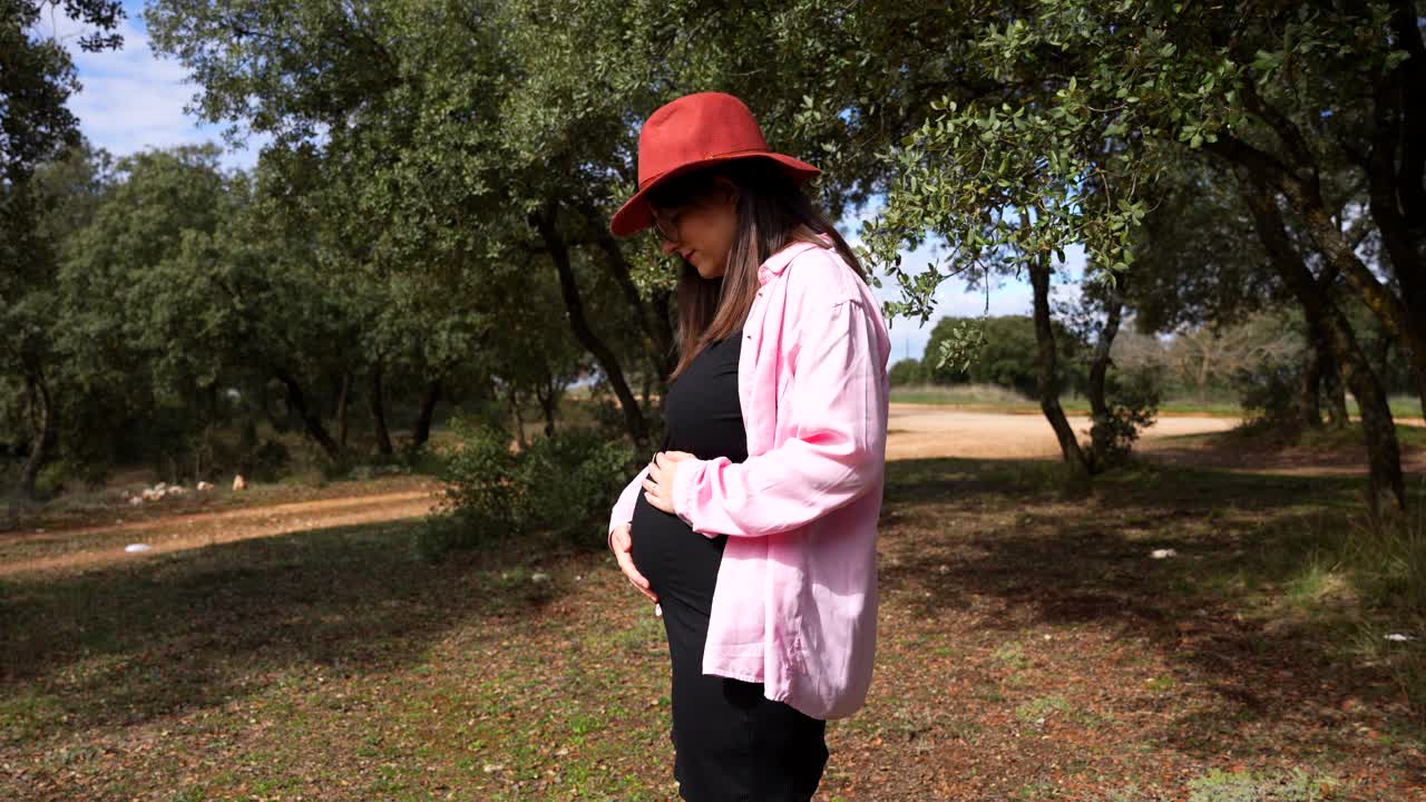 Pregnant woman in red hat gently touches her belly on sunny forest trail