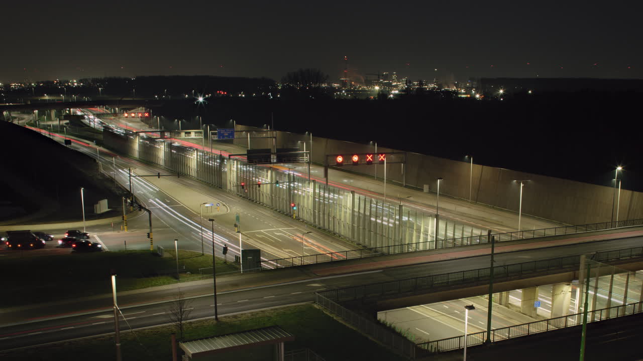 Traffic timelapse of a highway and intersecting roads transitioning into night, with streaking vehicle lights and an illuminated industrial skyline in the distance