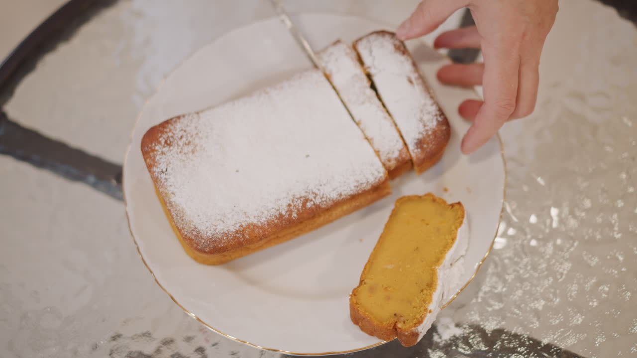 Closeup hands slicing pumpkin loaf dusted with powdered sugar on white plate at glass table, warm morning light, home baker serving golden spice bread with soft crumb, intimate cozy breakfast scene