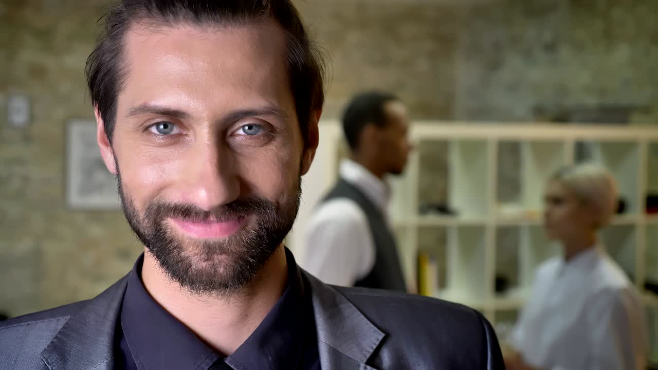 apuesto hombre de negocios confiado con barba está mirando a la cámara en la oficina, sonriendo, colegas están hablando en el fondo, concepto de comunicación