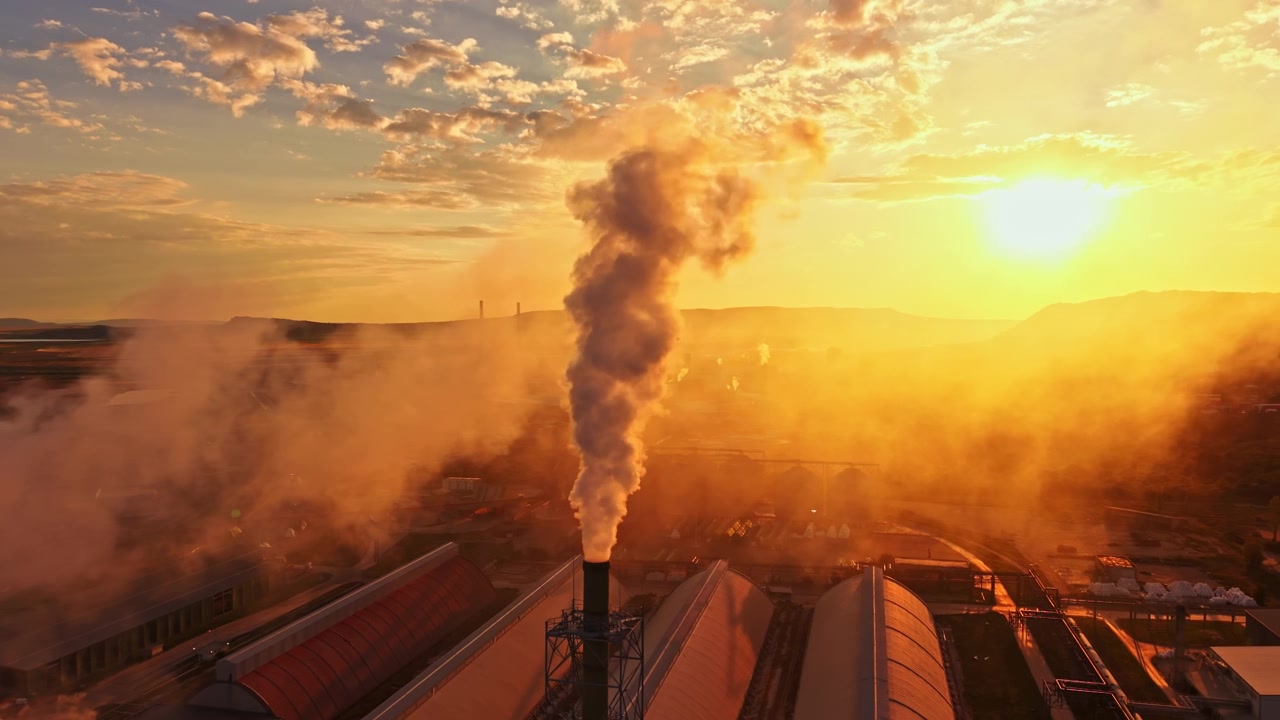 Smoke rises from a factory chimney during sunset with a view of the surrounding landscape and industrial buildings in the foreground